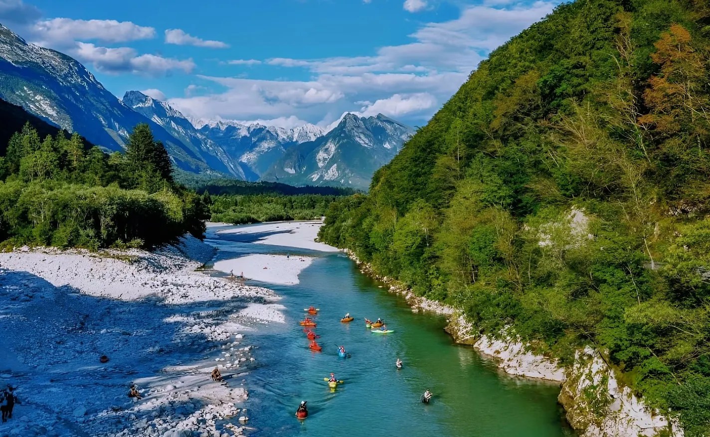 River with kayakers, flanked by green hills and distant mountains under a blue sky.